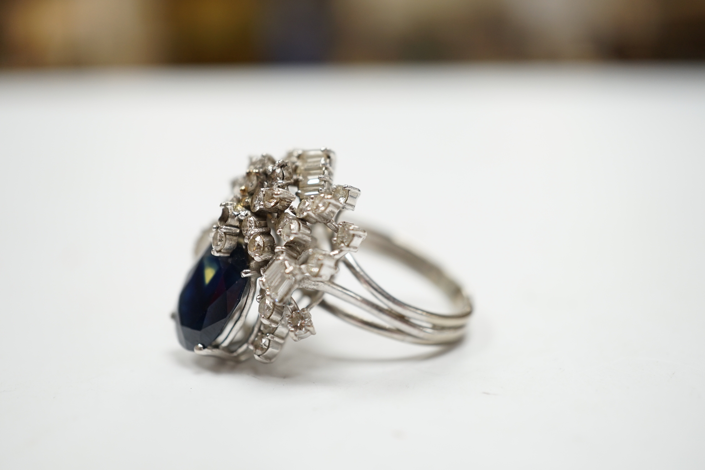 A mid to late 20th century 18k white metal, single stone oval cut sapphire and baguette and round cut diamond cluster set dress ring, size M/N, gross weight 10.3 grams.